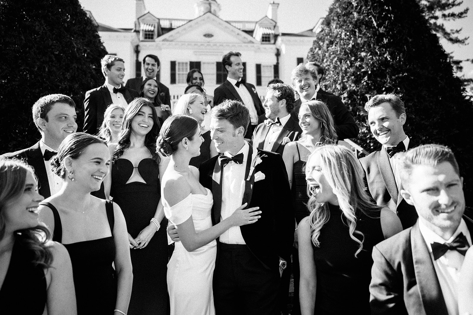 Black and white image of bride and groom surrounded by joyful wedding party on mansion steps
