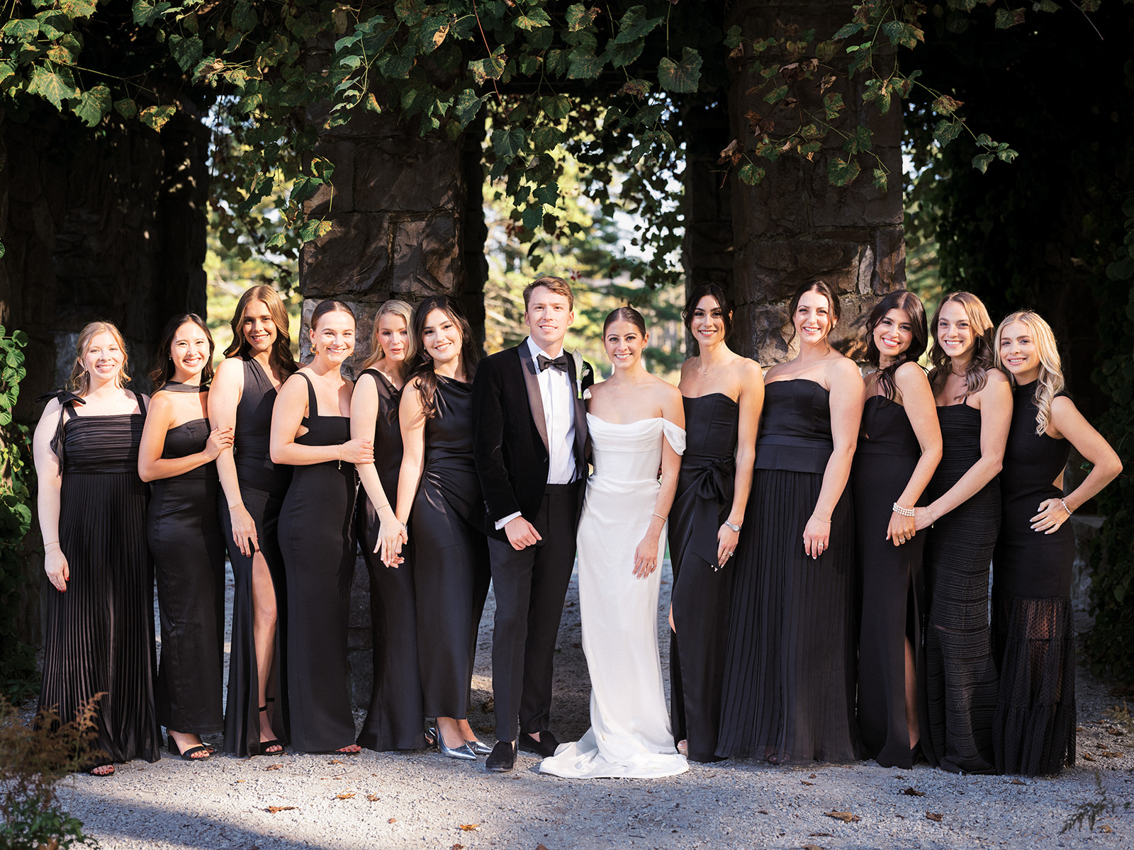 Bride and groom with bridesmaids in black dresses posed under vine-covered stone arch at The Mount
