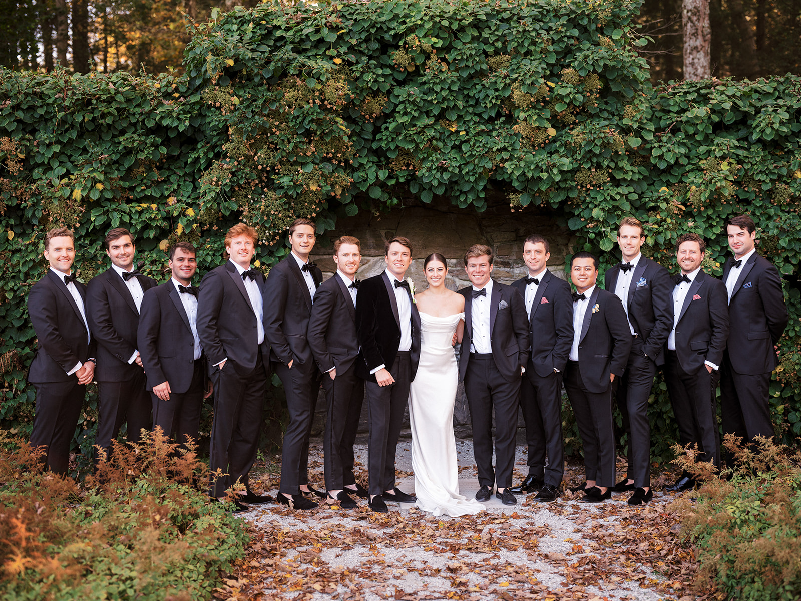Bride and groom with full groomsmen party posed in front of ivy-covered wall at Edith Wharton’s home