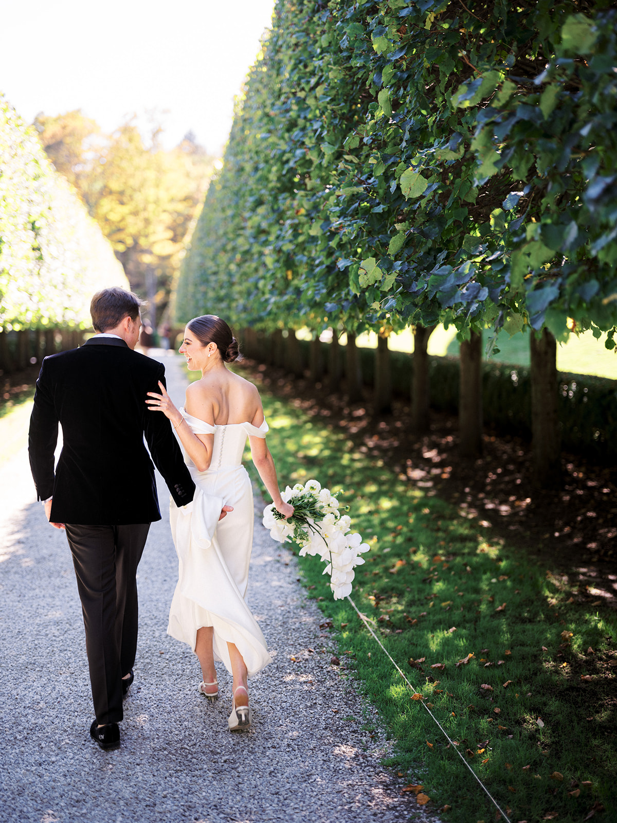 Bride and groom walking hand-in-hand through manicured tree-lined path at The Mount