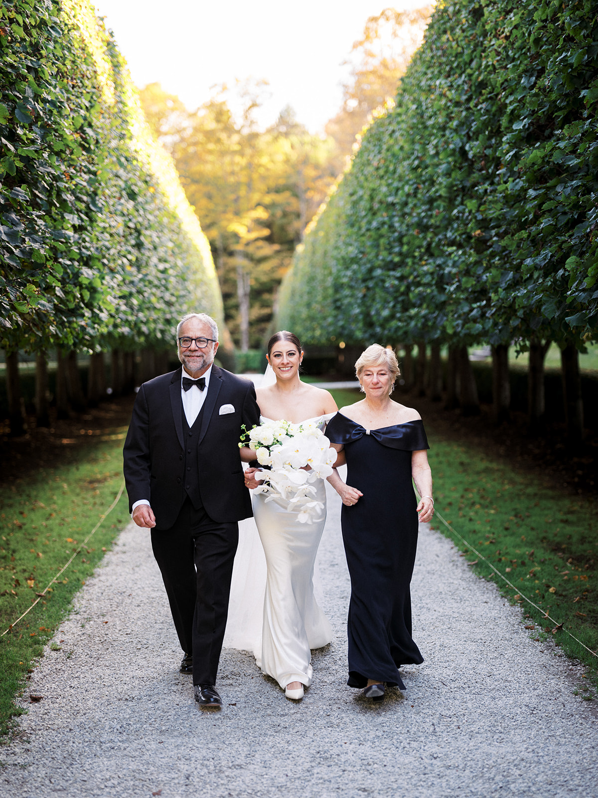 Bride walking down the aisle with her parents between tall, manicured hedges at The Mount.