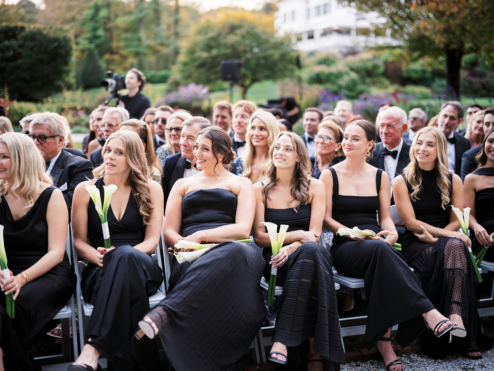 Bridesmaids in black dresses seated during the outdoor ceremony, each holding a single calla lily.