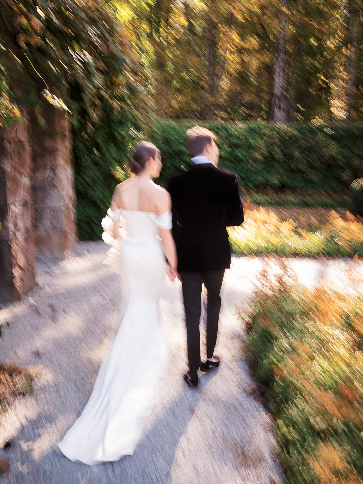Motion-blurred image of bride and groom walking hand-in-hand through wooded garden path
