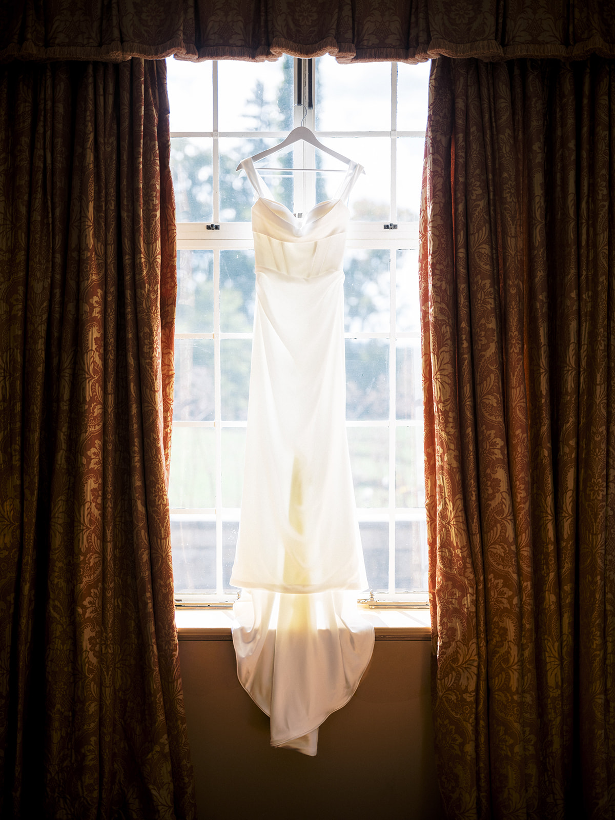 Wedding dress hanging in front of tall window with sunlight filtering through the fabric