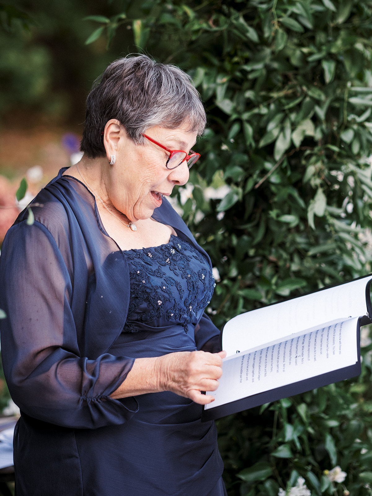 Wedding officiant reading from a book during the ceremony, wearing a navy dress with red glasses.